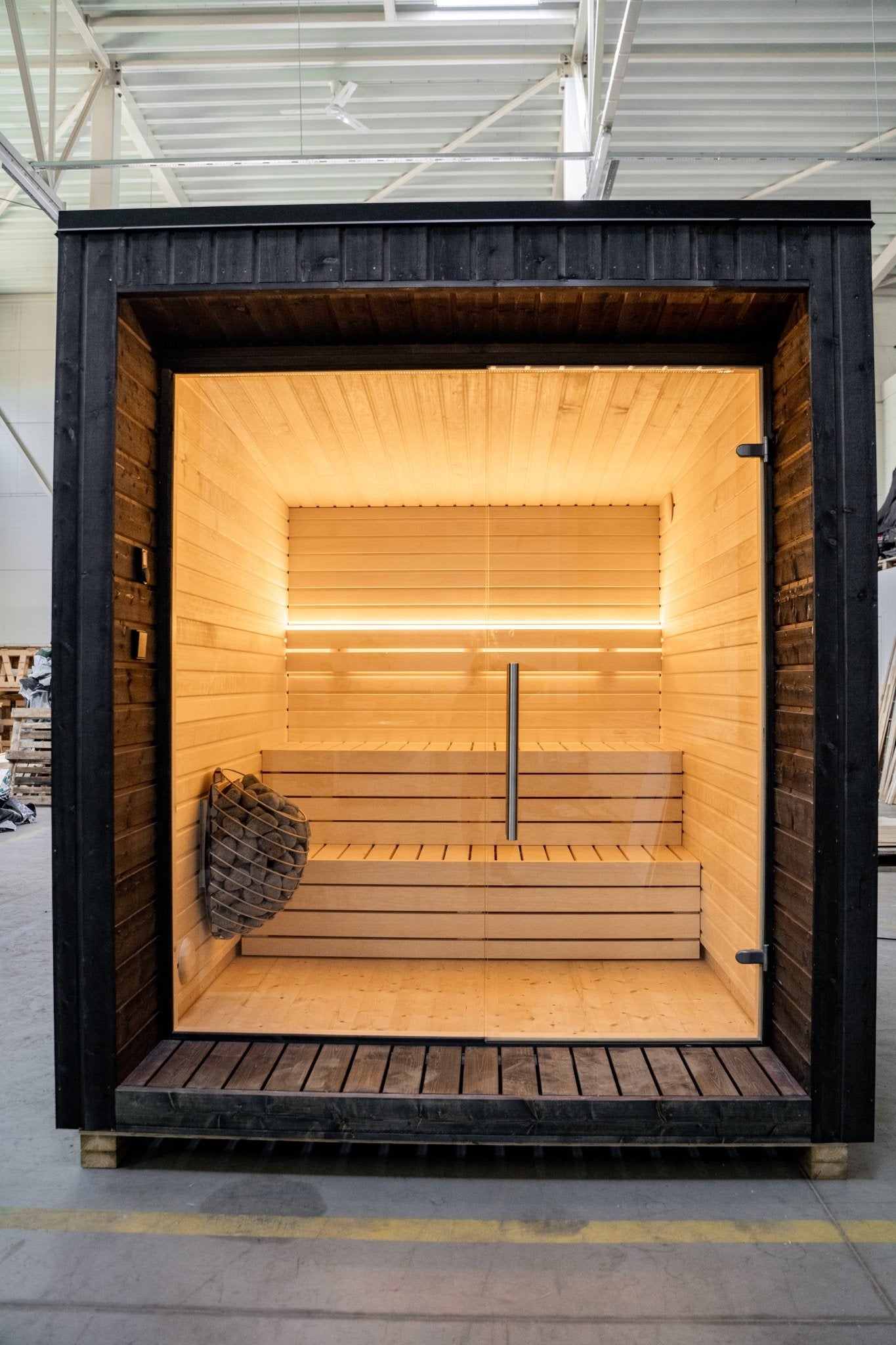 Sauna exterior Lumi con puerta de cristal y cesta de piedra.