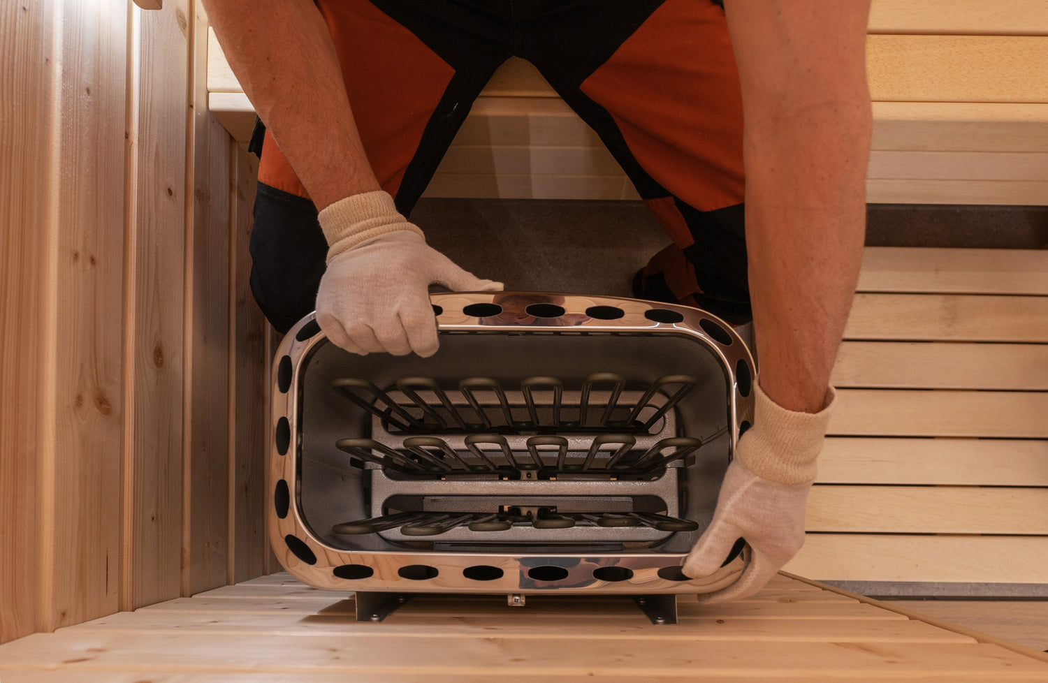 Person wearing orange and black work pants and white gloves is installing a stainless steel heater in a wooden room. The person is arranging the heater within a rectangular opening between wooden panels. The room appears to be part of a sauna.